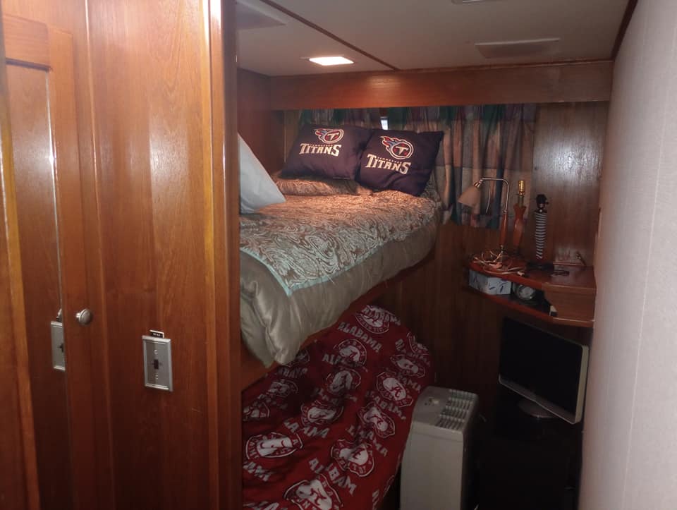 Interior view of a cozy bedroom featuring a queen-size bed with decorative bedding, two pillows with a sports team logo, and a lower bunk bed with red Alabama-themed bedding. The room includes wooden cabinetry and a small desk with a lamp.
