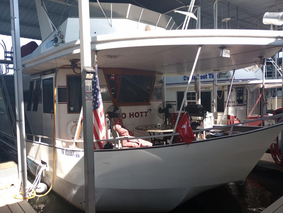 A 1990 Lazy Days 60' widebody houseboat docked at Cherokee Marina, featuring a white hull, a table on the deck, and an American flag displayed.