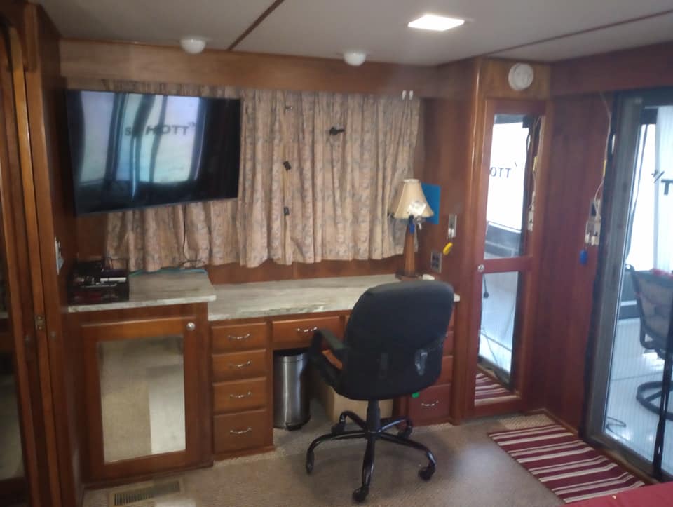 Interior view of a houseboat showing a desk area with a chair, a television mounted on the wall, and curtains. The room features wood paneling and a carpeted floor.