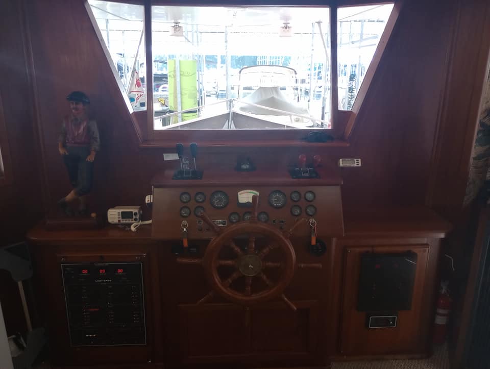 Interior view of the helm on a 1990 Lazy Days 60' houseboat, featuring a wooden steering wheel, control panel with gauges and levers, and a decorative figure.