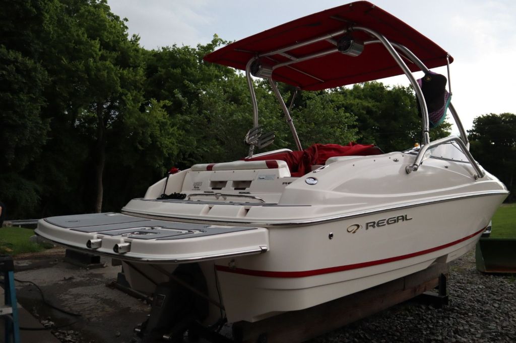 Side view of a 2007 Regal 2400 Bowrider boat, featuring a wakeboard tower and Bimini top, parked on a trailer in a wooded area.