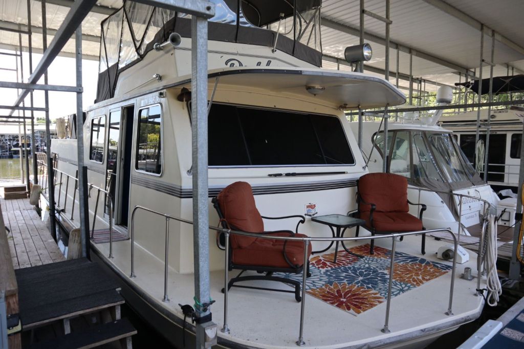A spacious houseboat featuring an outdoor seating area with two chairs and a table, located at Gallatin Marina. The boat is partially covered and surrounded by a dock.