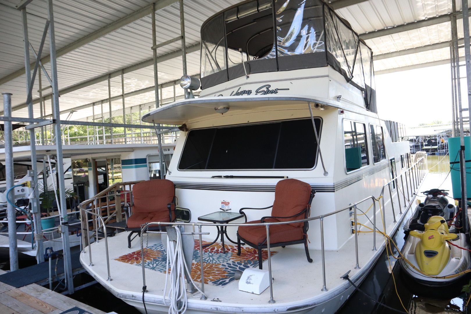 A houseboat with two brown chairs and a glass table on its deck, docked in a marina.