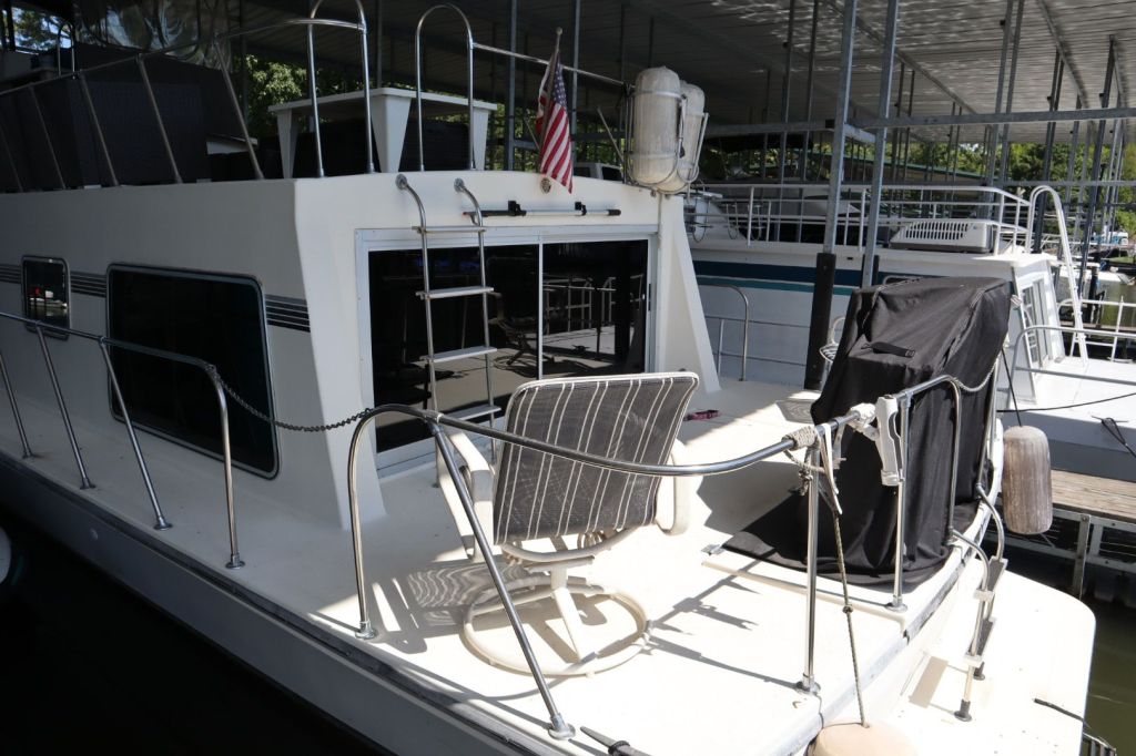 Exterior view of a 1986 Harbor Master 47' Widebody houseboat showcasing the deck with patio chairs, a ladder, and a view of the surrounding marina.