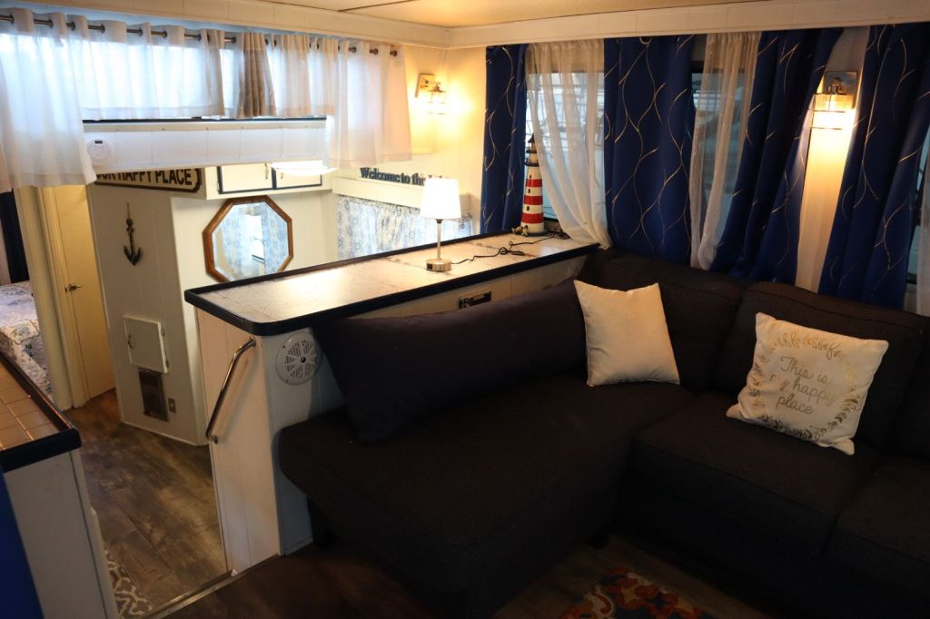Interior view of a cozy houseboat living space featuring a dark couch with decorative pillows, a lamp on a table, and blue curtains. There is a small kitchen area visible in the background along with a mirror and a nautical decor item.