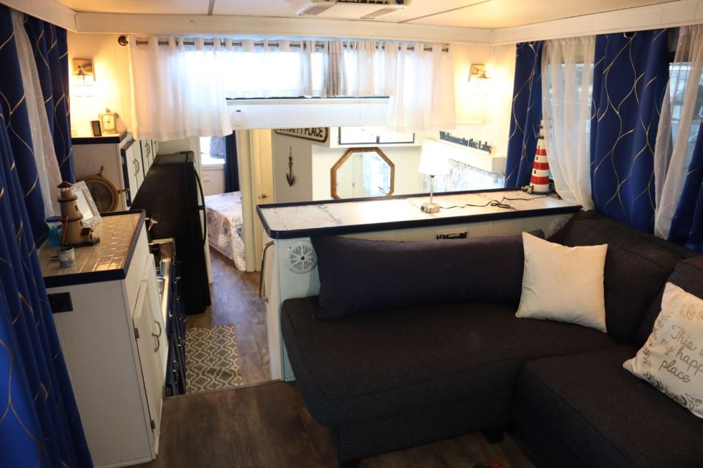 Interior view of a cozy houseboat featuring a dark gray sectional sofa, blue curtains, and a small galley kitchen with a decorative marine theme.