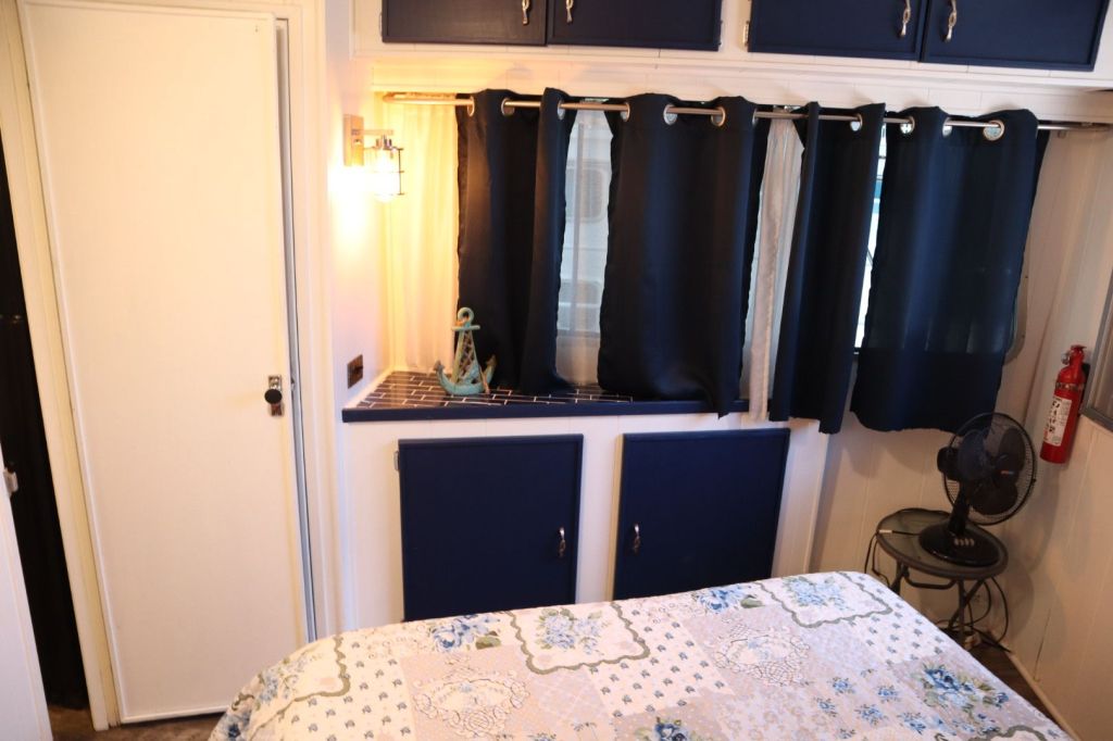 Interior view of a cozy bedroom in a houseboat featuring a floral patterned bedspread, blue curtains, wall-mounted lighting, and a small table fan.