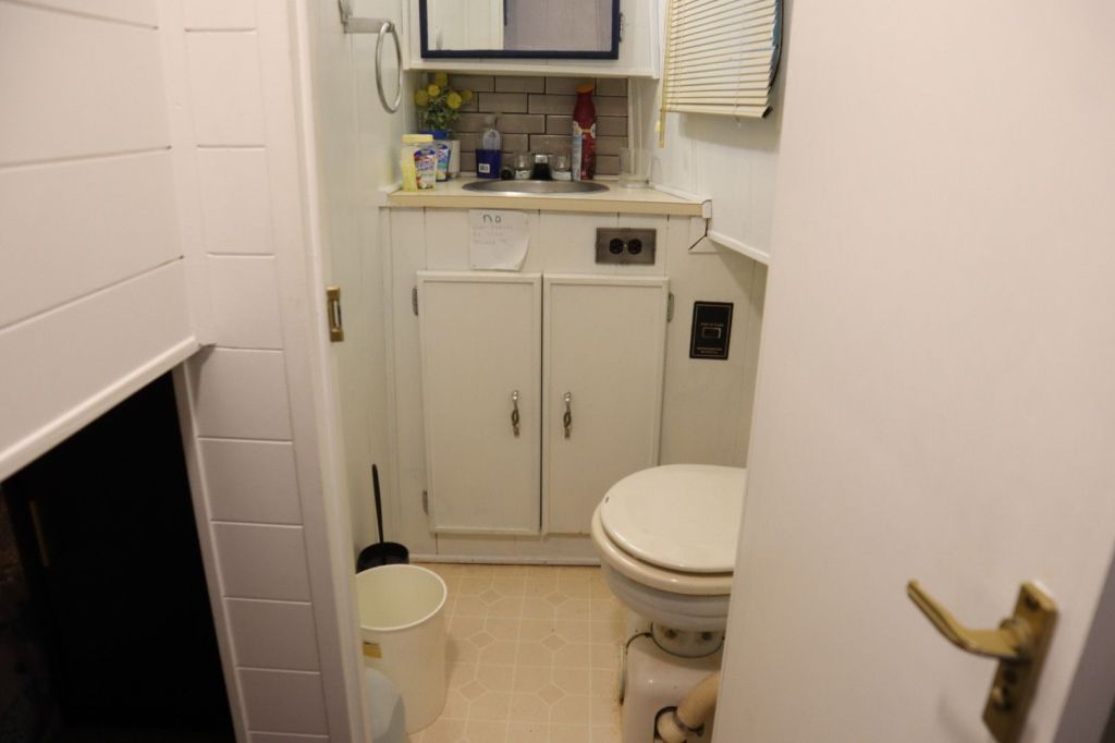 Interior view of a bathroom in a houseboat, featuring a toilet, sink with storage cabinets underneath, and a mirror. The walls are white, and a small window allows natural light.