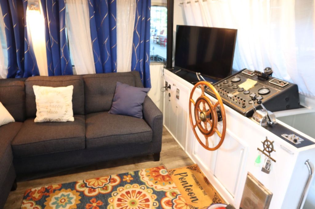 Interior view of a houseboat featuring a cozy seating area with a dark gray sofa and decorative pillows, accompanied by a steering wheel and navigation controls, a television, and colorful patterned rug.