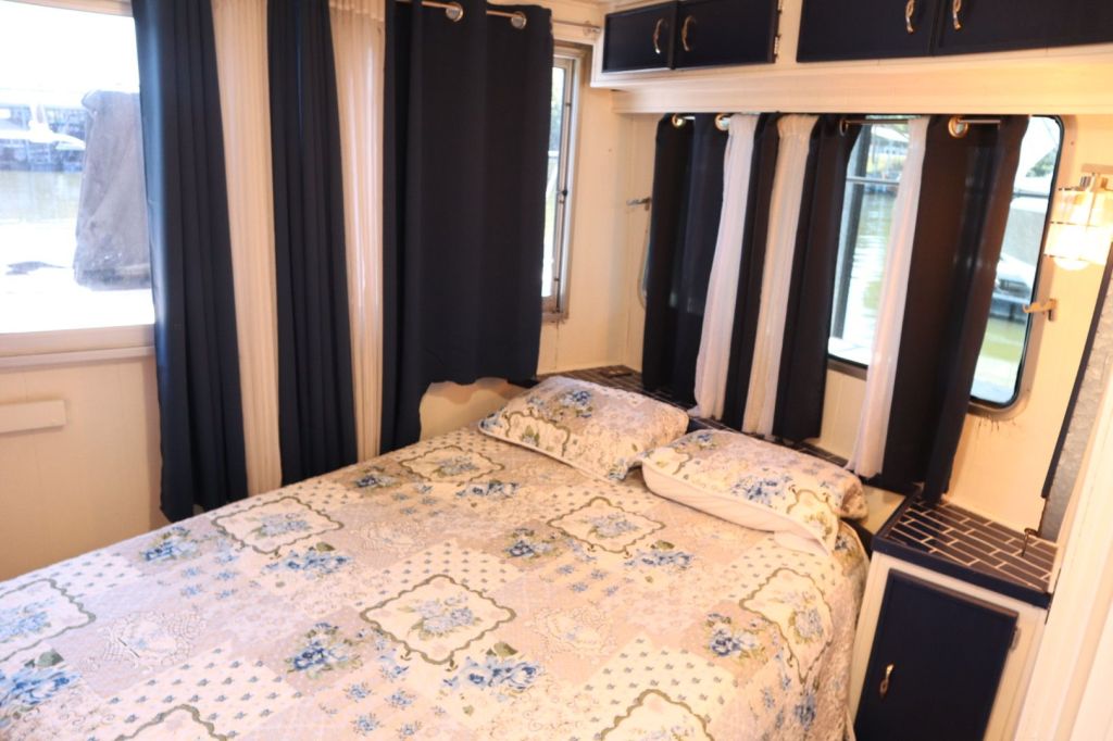 A cozy interior of a houseboat featuring a queen-sized bed with floral bedding, blue and white curtains, and windows revealing a view of the marina.
