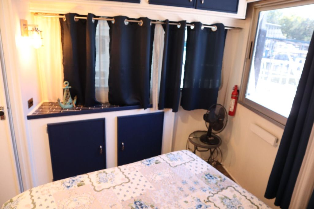Interior view of a cozy houseboat bedroom featuring a queen bed covered with a floral quilt, dark blue curtains, and decorative accents like an anchor. A small fan is visible near the window, along with storage cabinets.